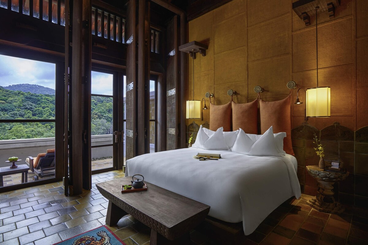 A hotel room with a large bed, wooden headboard, two bedside lamps, a small table with a tray, and a view of a lush landscape through an open door.