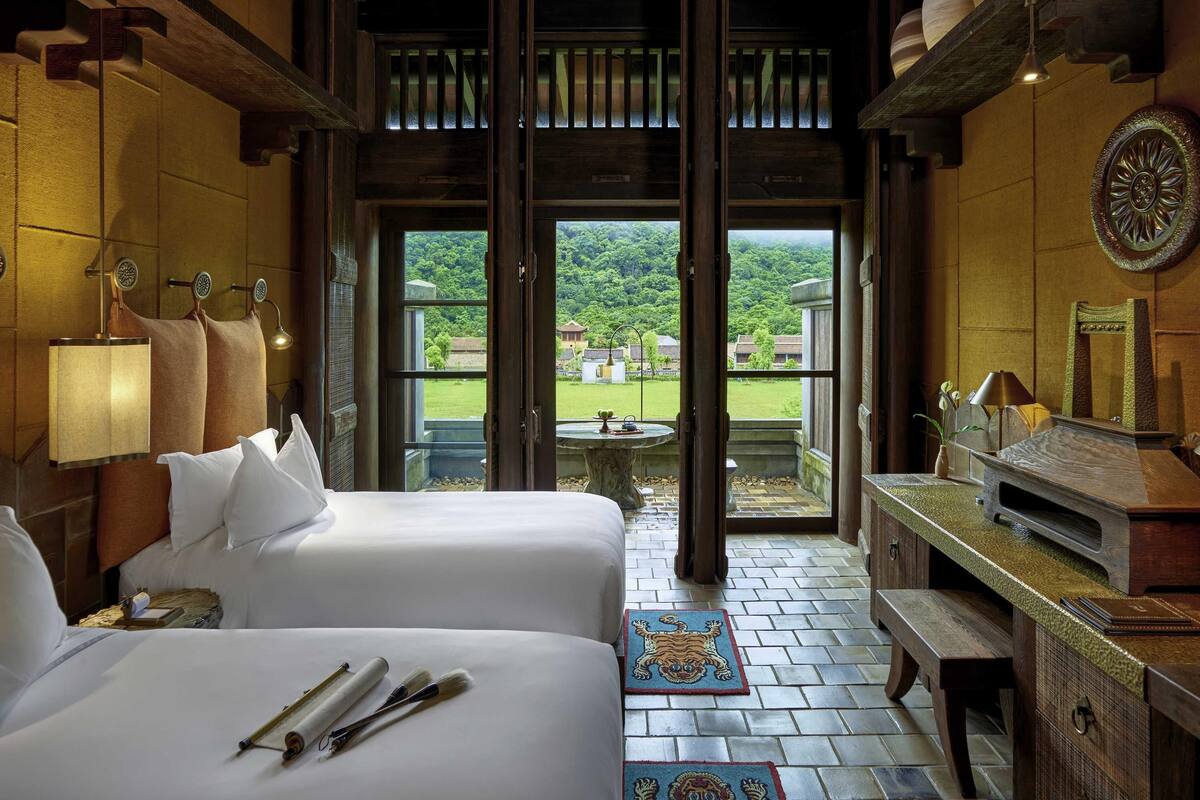 A hotel room with two beds, a desk, a TV, and a view of a green landscape.