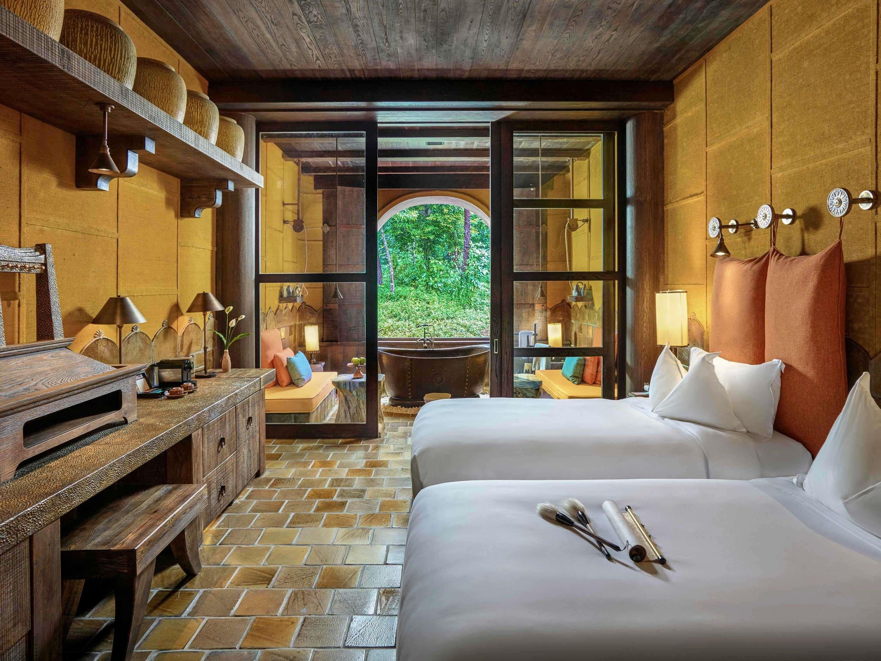 A room with two beds, a wooden desk, a large window, and a view of greenery outside.