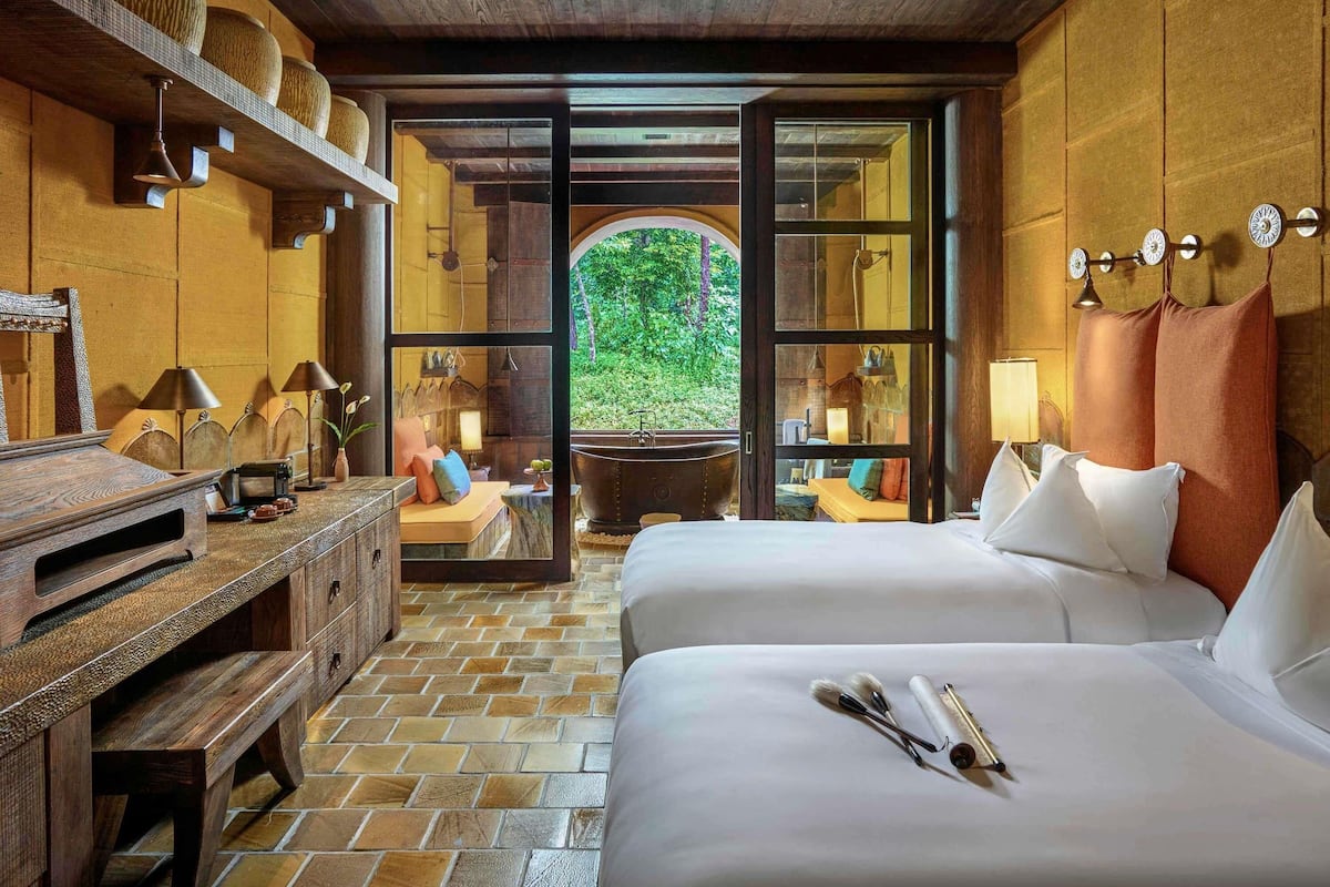A room with two beds, a wooden desk, a large window, and a view of greenery outside.