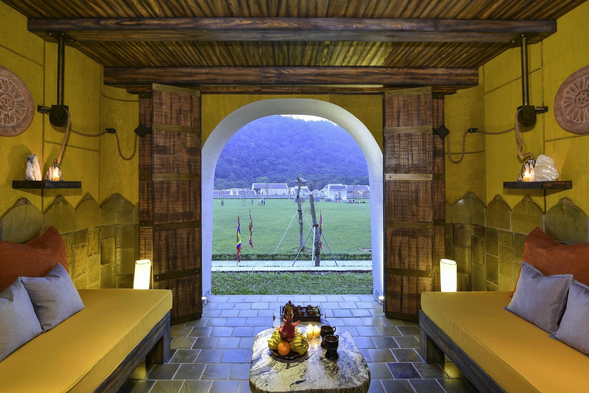 Una camera accogliente con una panca in pietra, un tavolo con frutta e candele, e vista su un campo e degli edifici attraverso un arco.