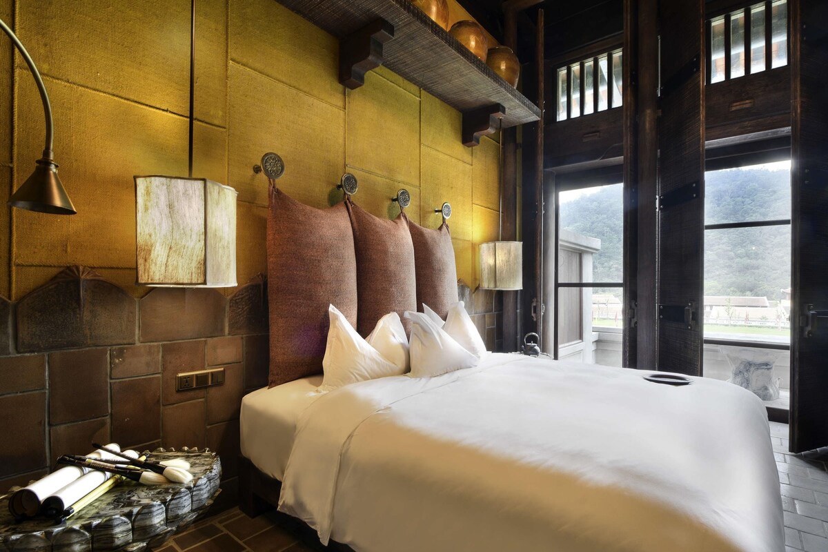 A large bed with white linens, a wooden headboard, and a brick wall. There are three wall-mounted lamps and a window with a view of greenery.