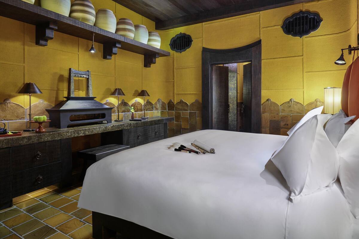Ein Hotelzimmer mit einem Bett, einem Schminktisch mit Spiegel, einer Ablage mit dekorativen Vasen und einer Tür zu einem weiteren Zimmer.
