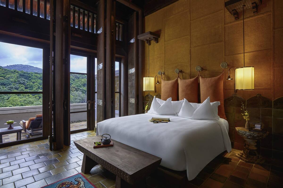 Ein Hotelzimmer mit einem großen Bett, einem hölzernen Kopfteil, zwei Nachttischlampen, einem kleinen Tisch mit Tablett und Blick auf eine üppige Landschaft durch eine offene Tür.
