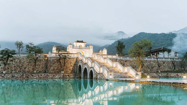 Un ponte in pietra tradizionale con il suo riflesso in uno specchio d'acqua tranquillo, che conduce a un edificio bianco con numerosi archi e uno sfondo montuoso.