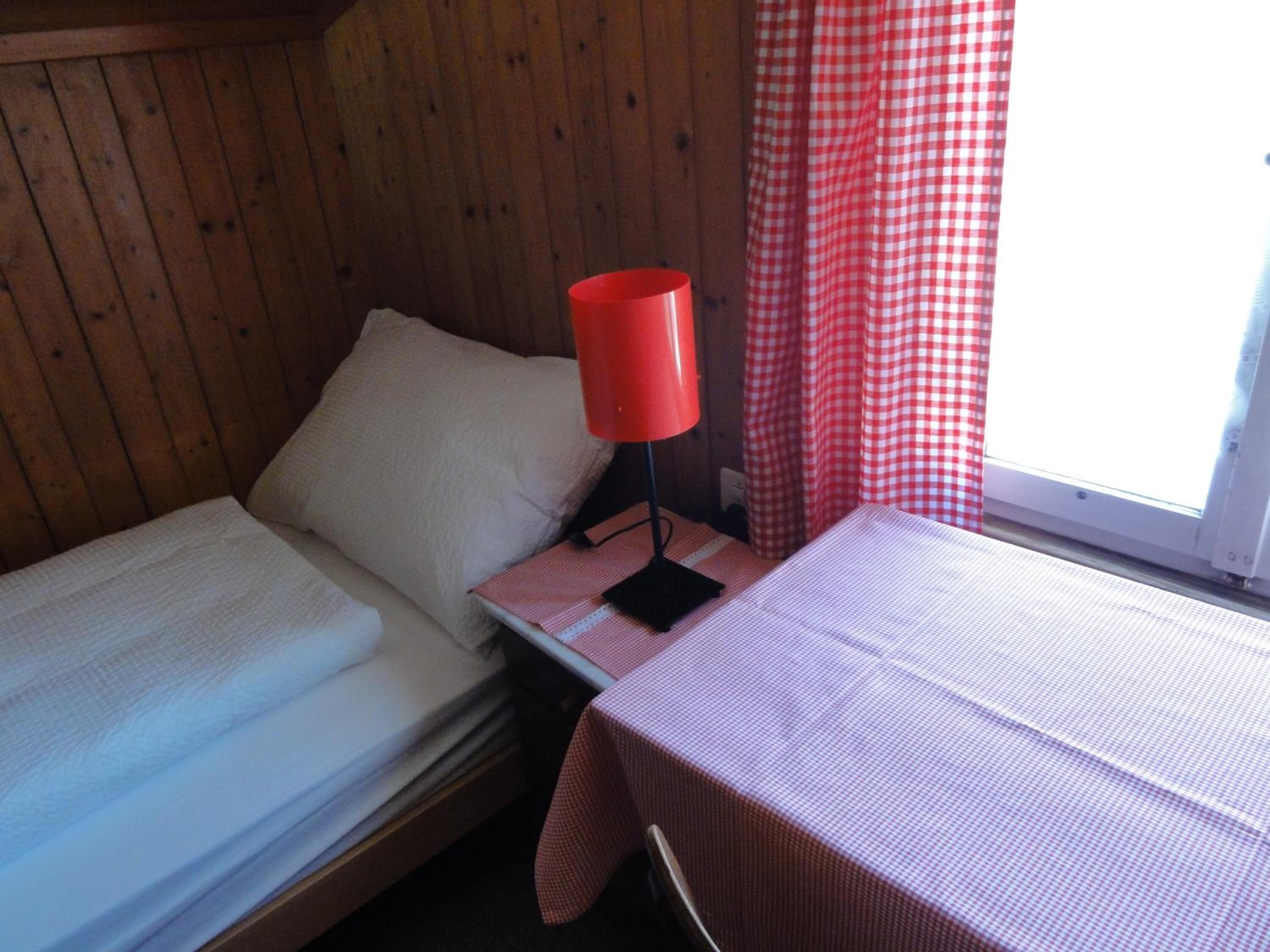 A small, cozy room with two single beds, a red lamp on a nightstand, and a window with red and white checkered curtains.
