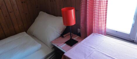 A small, cozy room with two single beds, a red lamp on a nightstand, and a window with red and white checkered curtains.