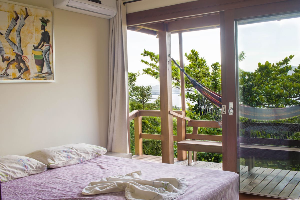 Een slaapkamer met een bed, een schilderij en een balkon met uitzicht op groen en een wateroppervlak.