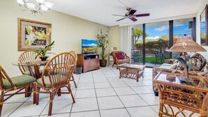 Un vaste espace de vie comprenant une table à manger, des chaises en osier, un canapé et un téléviseur. Un ventilateur de plafond est installé et de grandes fenêtres offrent une vue sur l’extérieur.