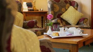 Una habitación acogedora con un sillón floral, una mesa de madera con una bandeja de té y galletas, y un jarrón con una sola rosa.