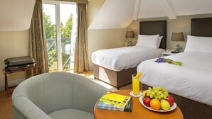 Habitación de hotel con dos camas, un balcón, una silla, una mesa con fruta y un libro, y una maleta.