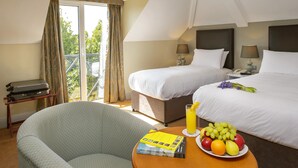 Habitación de hotel con dos camas, un balcón, una silla, una mesa con fruta y un libro, y una maleta.