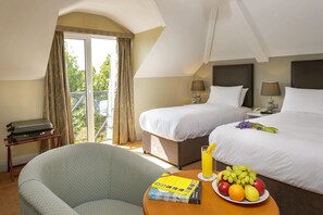 Une chambre d’hôtel avec deux lits, un balcon, une chaise, une table avec des fruits et un livre, et une valise.