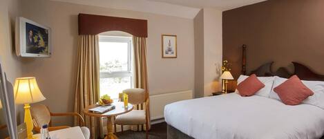 Een hotelkamer met een bed, nachtkastje, lamp, stoel, raam en een klein tafeltje met een lamp en een fles.