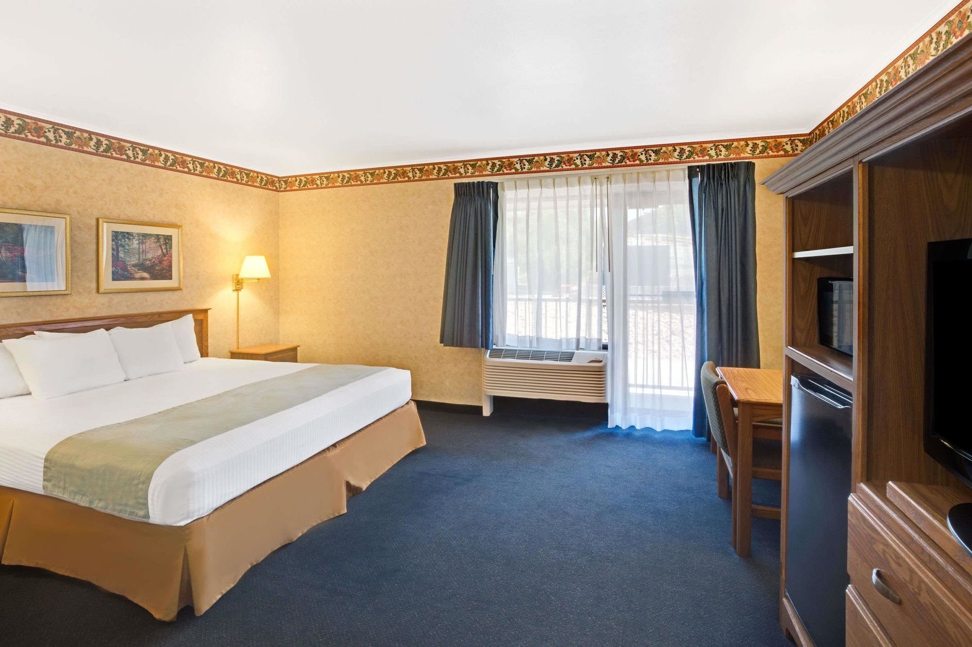 A hotel room with a bed, a desk with a chair, a television, and a window with curtains.