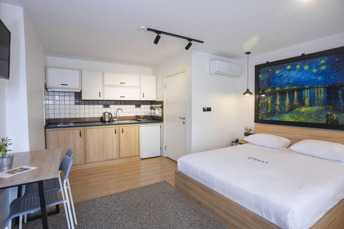 A modern hotel room with a wooden bed, a dining area with a table and chairs, a kitchenette, and a large painting of a night scene on the wall.