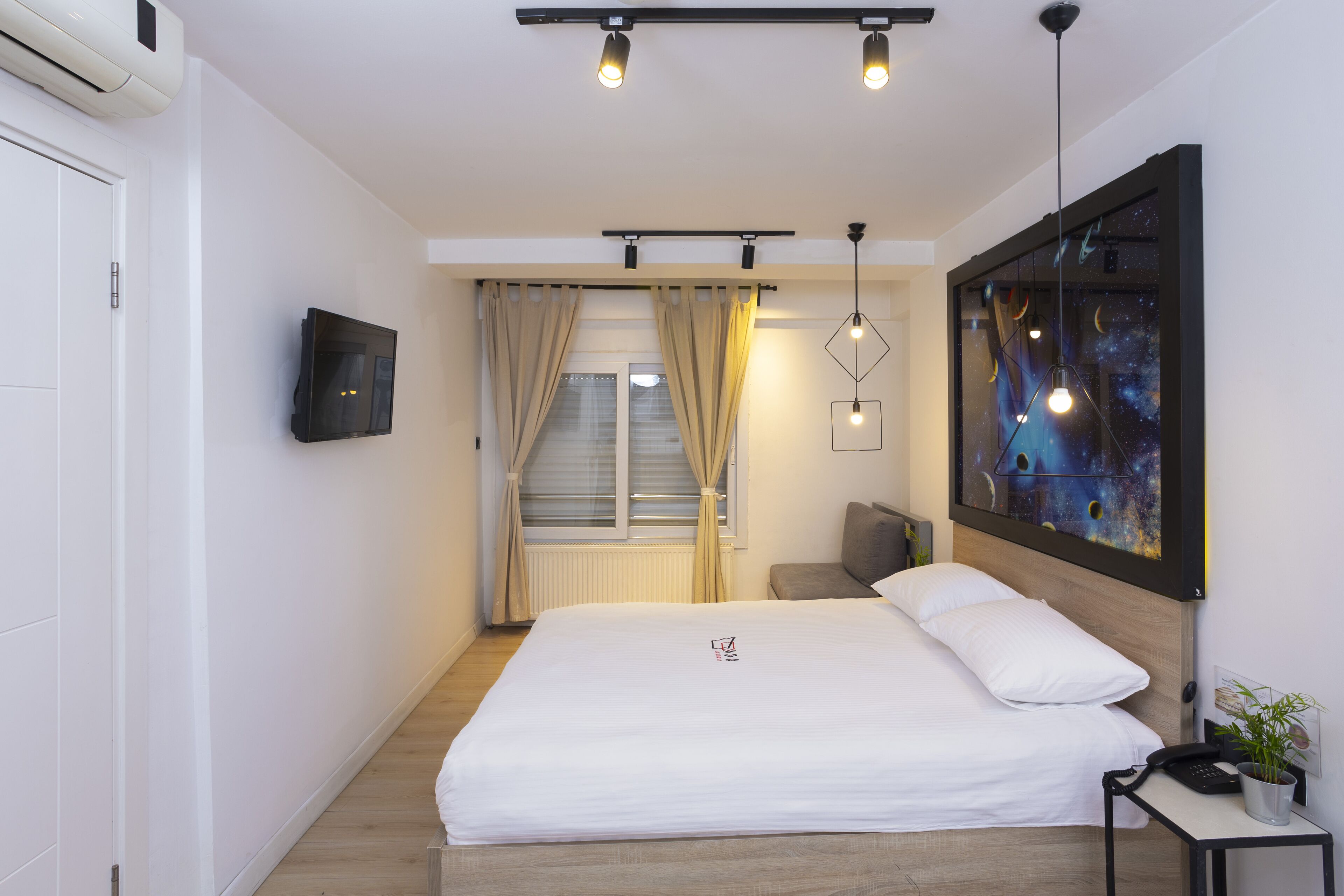 A modern hotel room with a large bed, a flat-screen TV, a desk with a phone, and a painting of a galaxy on the wall.