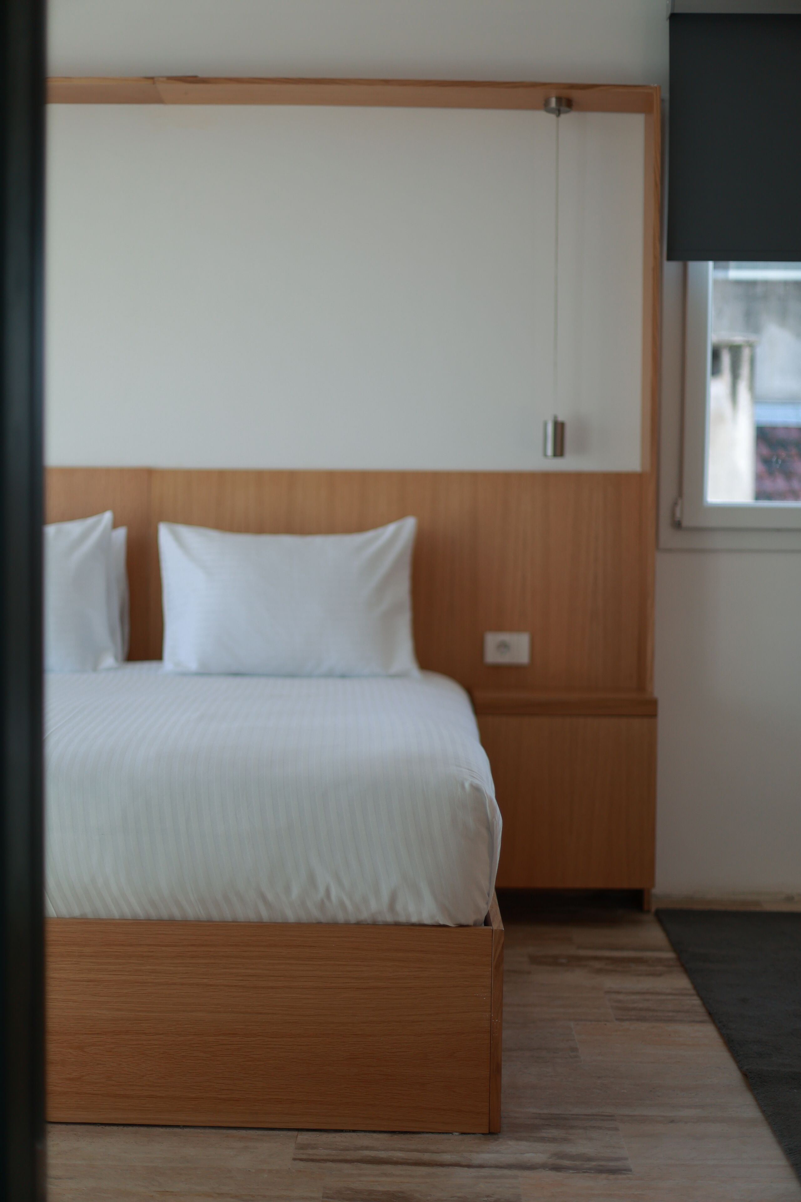 Une chambre à coucher avec une tête de lit en bois, un lit recouvert de draps blancs, une table de chevet et une fenêtre donnant sur des immeubles.