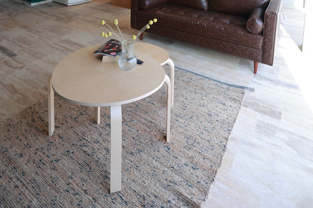 A modern living area with a brown leather sofa, a round table with a vase, and a kitchen in the background.