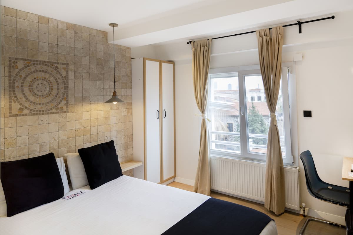 A hotel room with a bed, a desk, a window with curtains, and a wall with a mosaic pattern.