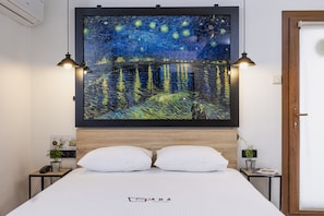 A hotel room with a bed, two bedside tables, a nightstand, a wall-mounted air conditioner, and a framed painting above the bed. - Ada Apart (Izmir)