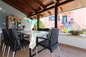 Un patio al aire libre con una mesa y sillas, techo de madera y un edificio de fachada rosa al fondo.