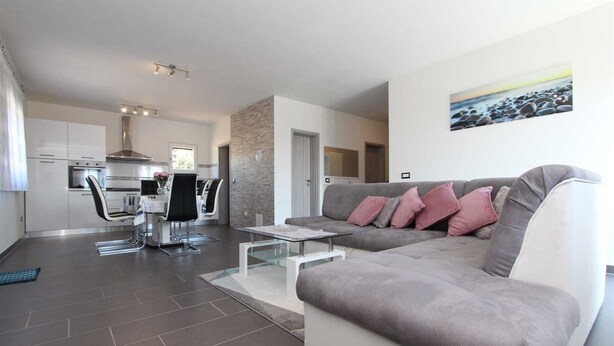A modern living room with a grey sofa, a coffee table, and a dining area with chairs and a kitchen in the background. - MONDELACO Three-Bedroom Apartment (Rovinj)