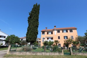 Un edificio de varios pisos con techo de tejas rojas, cerca verde y un árbol alto al frente.
