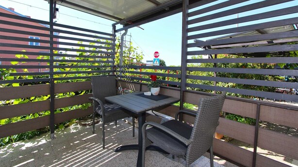 Een overdekt buitenzitgedeelte met een tafel en stoelen, omgeven door een metalen hek met horizontale latten.