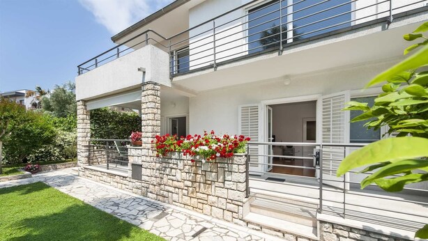 A two-story house with a stone facade, a balcony with metal railings, and a flower box with red flowers.