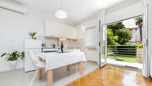 A modern kitchen with white cabinets, a dining table with a white tablecloth, and a view of a green garden through an open door.
