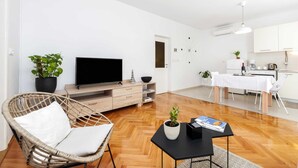 A modern living room with a flat-screen TV, a wicker chair, a dining area with a white table, and a kitchen with a coffee machine.