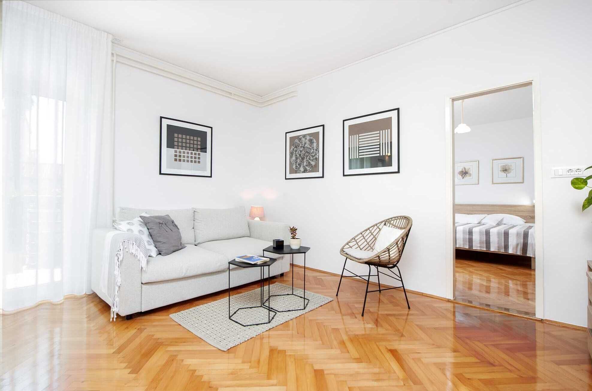 A modern living room with a sofa, a chair, a coffee table, and a bed in the background. There are three framed pictures on the wall.