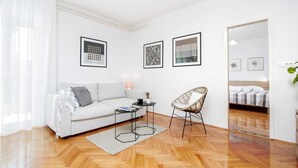 A modern living room with a sofa, a chair, a coffee table, and a bed in the background. There are three framed pictures on the wall.