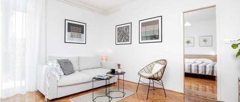 A modern living room with a sofa, a chair, a coffee table, and a bed in the background. There are three framed pictures on the wall.