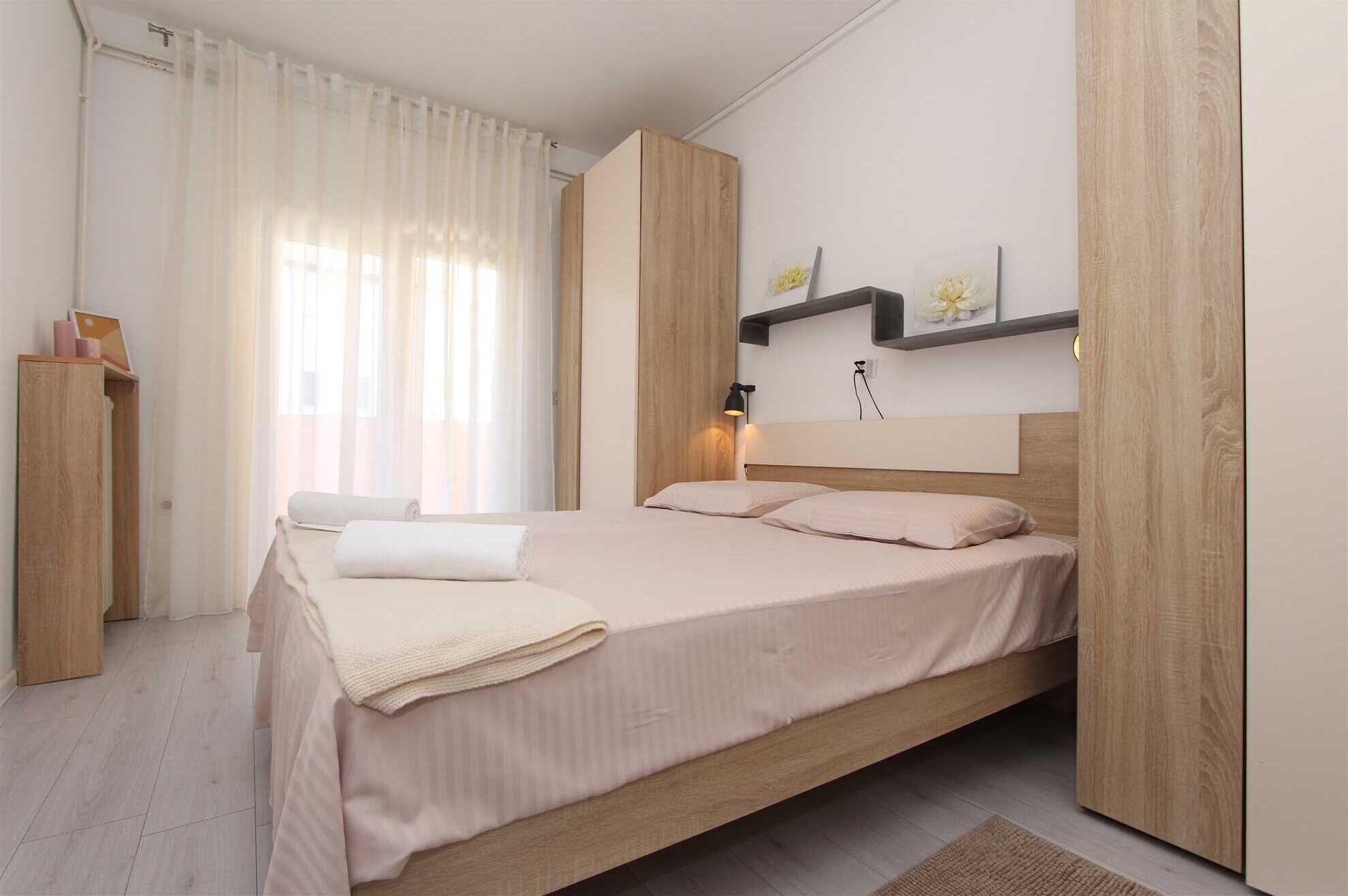 A bedroom with a bed, a nightstand, a wooden wardrobe, a window with sheer curtains, and a wall-mounted shelf with decorative items.
