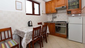 Une petite cuisine avec des meubles en bois, une cuisinière, un évier et un réfrigérateur. Il y a une fenêtre au-dessus de l’évier et une chaise avec un coussin à rayures.