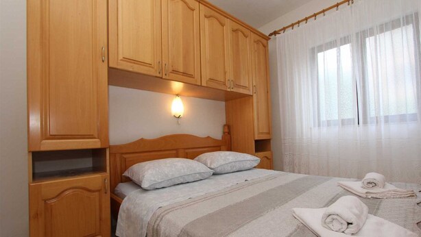 Une chambre avec des armoires en bois, un lit doté d’une tête de lit, un luminaire fixé au mur et une fenêtre ornée de rideaux légers.