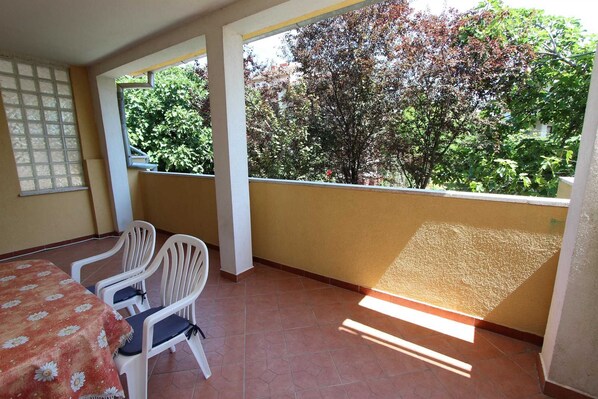 Un balcón con suelo de baldosas, dos sillas blancas, una mesa con mantel floreado y vistas a árboles y follaje.