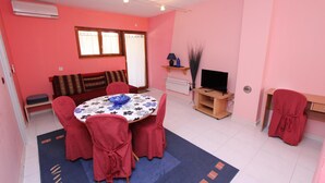 Ein Essbereich mit einem runden Tisch, roten Stühlen und einer Tischdecke mit Blumenmuster. Es gibt eine Couch und eine TV-Konsole mit einem Flachbildfernseher.