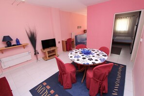 Una habitación con decoración rosa, una mesa redonda, sillas rojas, un televisor, un radiador y una puerta que da a un dormitorio.