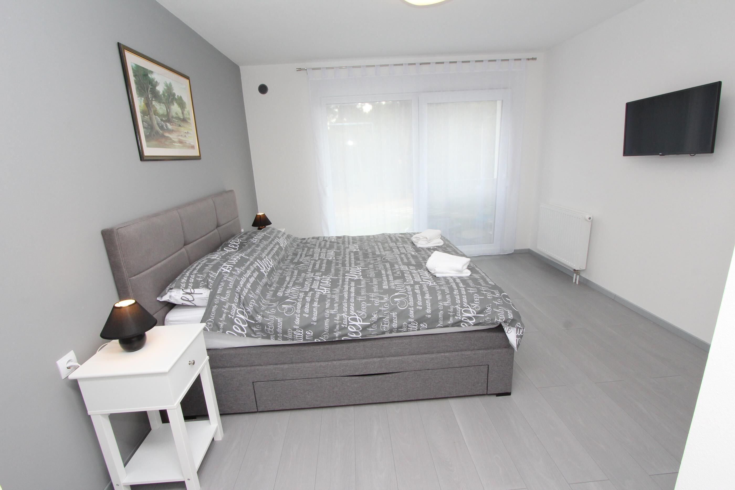 A modern bedroom with a grey bed, white bedside table, and a mounted TV.