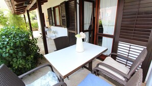 Un patio al aire libre con una mesa y sillas de mimbre, un jarrón blanco con flores y vistas a un jardín.