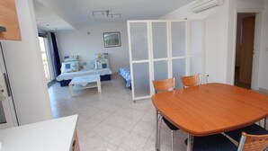 Ein Zimmer mit Holztisch, zwei Stühlen, einem Bett mit blau-weißer Bettdecke, einem weißen Nachttisch und einem weißen Kleiderschrank mit Milchglas-Türen.