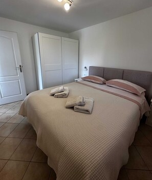 Una cama bien hecha con una colcha color beige y almohadas, un armario blanco y un piso de baldosas.