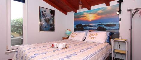 Un dormitorio con una cama, un globo terráqueo, un cuadro de un atardecer y una ventana con vistas a la vegetación.