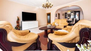 Ein Wohnzimmer mit Ledersesseln, einem Flachbildfernseher, einer Chandelüster und einem durch einen Bogen sichtbaren Essbereich.
