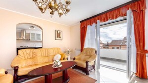 Ein Wohnzimmer mit Sofa, Sesseln, einem Couchtisch, einer Chandelüster und Blick auf die Stadt durch große Fenster mit orangefarbenen Vorhängen.