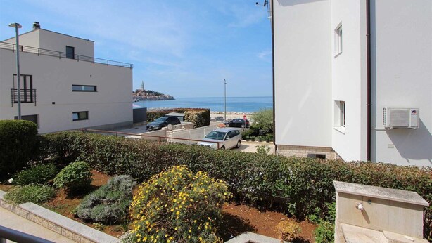 A coastal building with a view of the sea, a parking area, and a garden with yellow flowers.