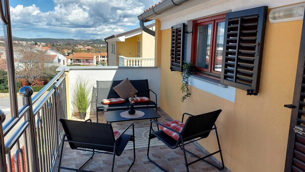 A balcony with two black chairs, a small table, and a sofa with red and white cushions. There is a window with black shutters and a potted plant on the wall. - MATOSEVIC One-Bedroom Apartment with Balcony 1 (Rovinj)